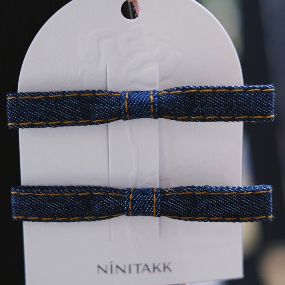 A pair of dark denim alligator clips, each adorned with a slim, stylish bow, perfect for adding a modern and casual touch to any hairstyle.