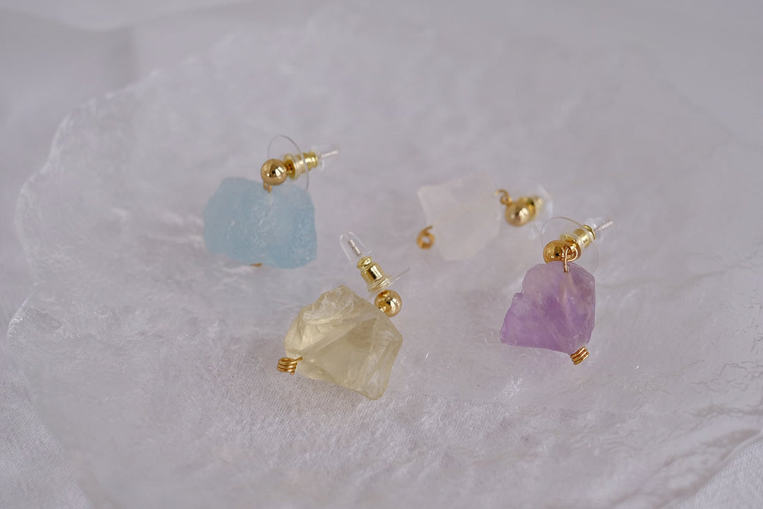 The earrings are arranged on a white textured surface, showcasing the vibrant colors and raw, natural shapes of the crystals.