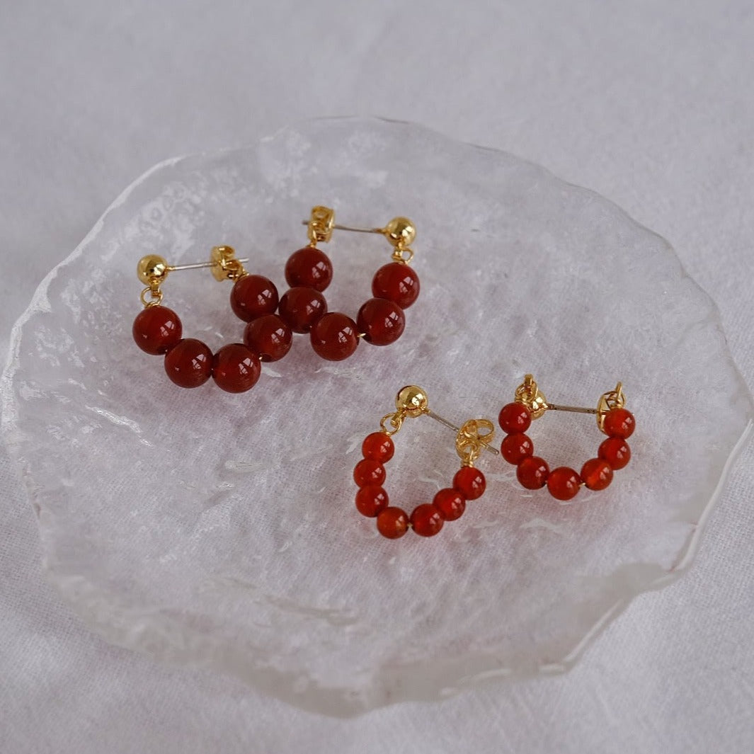 A close-up shot of both sizes of the red onyx hoop earrings on a clear dish, highlighting their vibrant red color and gold accents.