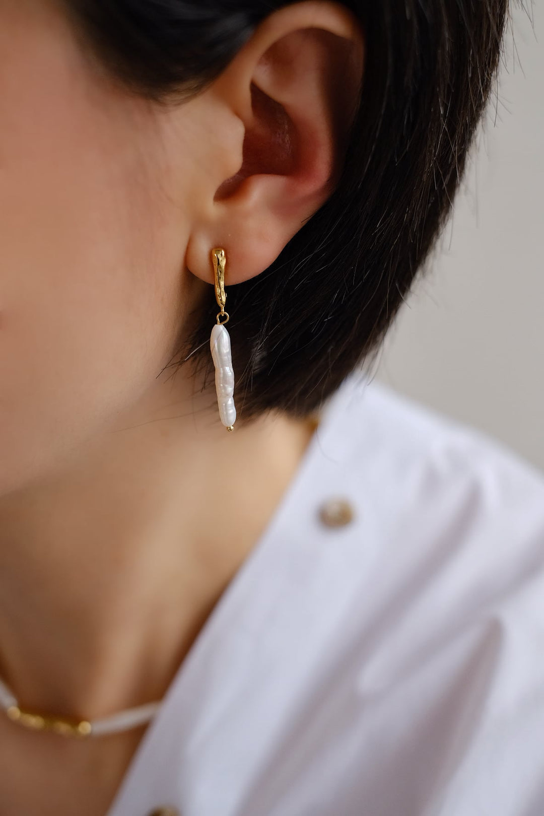 The earrings showcase a delicate composition, with a long, thin golden cylinder connected to a longer baroque pearl, offering a handcrafted aesthetic.