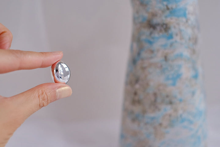 A close-up of a hand holding one earring, showcasing the smooth, reflective surface and oval shape.