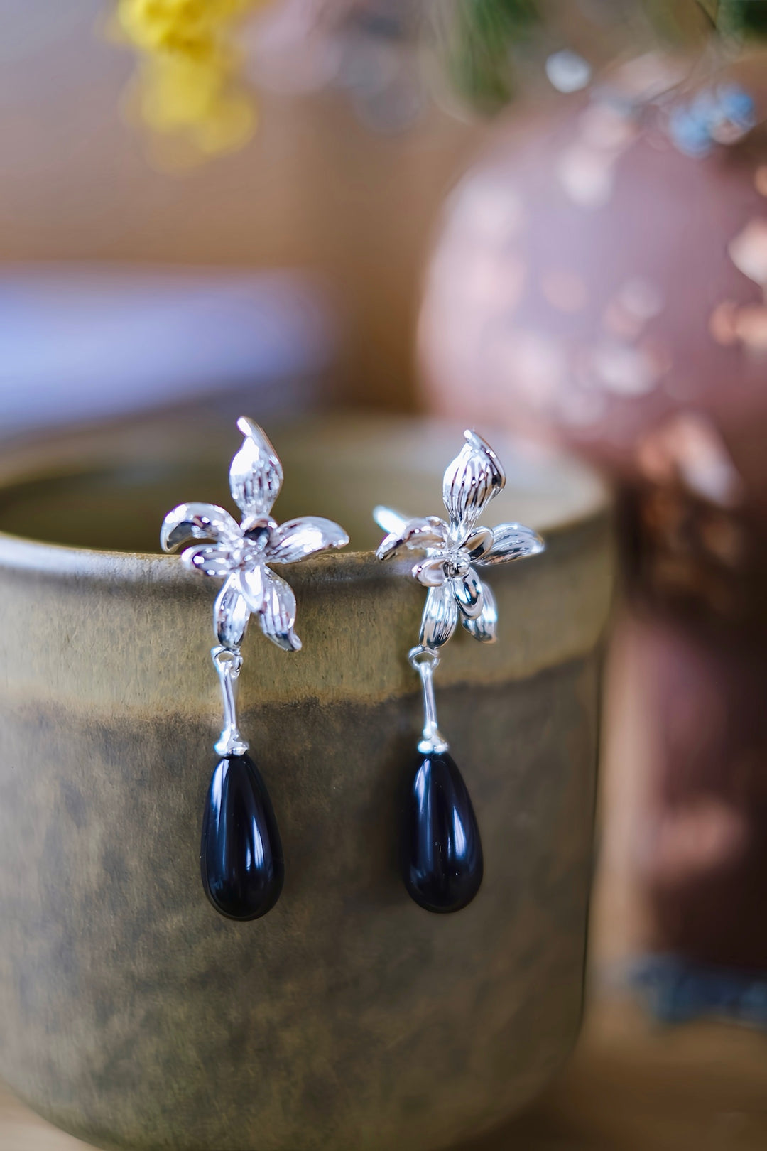 Close-up of the Daffodil earrings hanging from the rim of a ceramic cup, highlighting the intricate silver-plated brass flower design and the elegant black teardrop dangle.